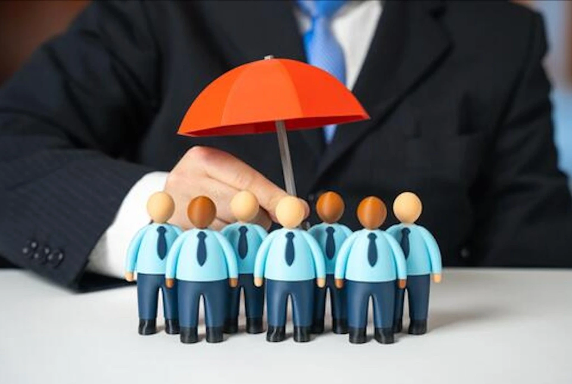 Figurines de collaborateurs en costume sur un bureau blanc, protégés par un parapluie rouge tenu par une personne en costume professionnel — métaphore de la couverture multirisque entreprise